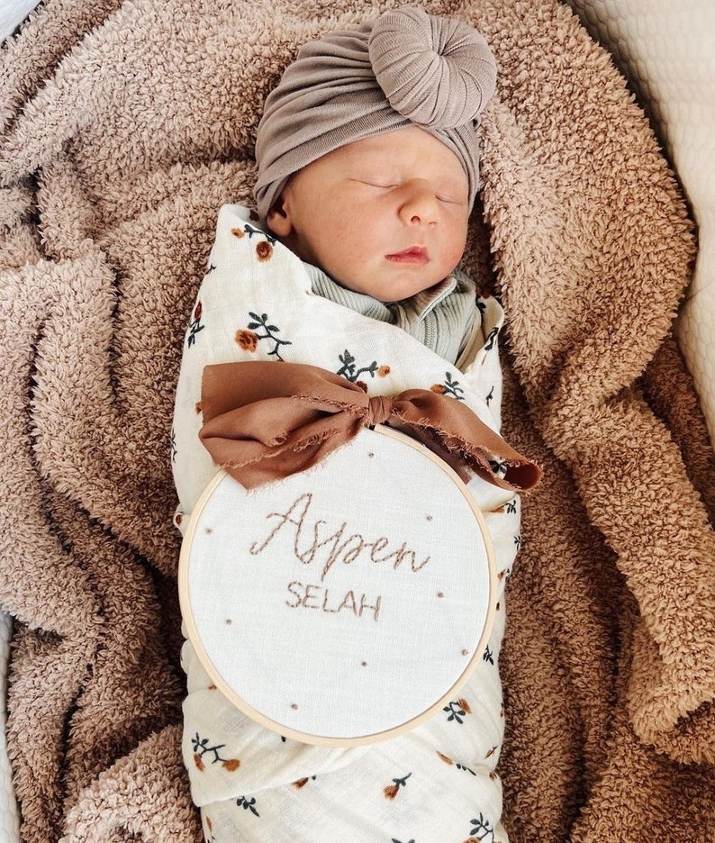Puede incluir: Un beb&eacute; reci&eacute;n nacido con un turbante marr&oacute;n y un arrullo blanco con estampado floral. El beb&eacute; est&aacute; acostado sobre una manta peluda marr&oacute;n. Un aro de bordado de madera con tela blanca y el texto "Aspen Selah" est&aacute; delante del beb&eacute;.