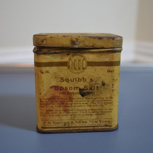 Peut inclure: Une boîte en métal jaune avec une étiquette blanche qui indique "Squibb's Epsom Salt (True Magnesium Sulphate)". L'étiquette comprend également des instructions d'utilisation et le nom et l'adresse de la société : "E.R. Squibb & Sons, New York".