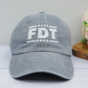 Puede incluir: Gorra de béisbol gris con las letras blancas "FDT" bordadas en la parte delantera. Las letras están entre líneas y estrellas blancas. La gorra está hecha de un material suave y tiene una visera curva.