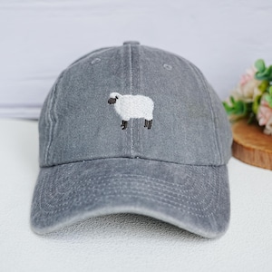May include: A gray baseball cap with a white embroidered sheep design on the front. The cap has a curved brim and an adjustable strap at the back. The sheep is depicted with a white body, black legs, and a black head.