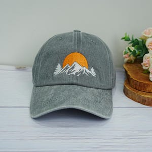 Puede incluir: Gorra de béisbol gris con un diseño bordado de montaña y sol. La gorra presenta una visera curva y una construcción clásica de seis paneles. La escena de la montaña está representada en hilo blanco y naranja, creando un contraste llamativo.