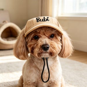 Peut inclure: Une casquette de baseball beige pour chien avec le nom Bubi brodé en écriture noire sur le panneau avant. La casquette comprend une mentonnière noire réglable avec un bloqueur de cordon, conçue pour maintenir le chapeau sur la tête.