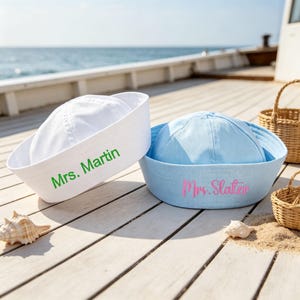 Może przedstawiać: Dwie czapki marynarskie, jedna biała z zielonym napisem Mrs. Martin i jedna jasnoniebieska z różowym napisem Mrs. Slater. Czapki mają zaokrągloną główkę i szeroki, wywinięty rondo, umieszczone na drewnianym pokładzie obok małych muszli.