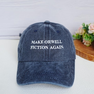 Peut inclure: Casquette de baseball bleu denim avec l'inscription "MAKE ORWELL FICTION AGAIN" brodée en blanc. La casquette a une visière incurvée et un bouton sur le dessus. En arrière-plan, une surface en bois et des fleurs.