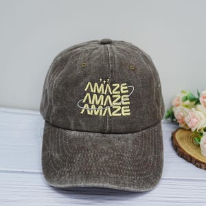 May include: A dark brown, washed-out cotton baseball cap featuring the word "AMAZE" embroidered three times in yellow thread, stacked vertically with a thin, curved silver line encircling the text and three small stars above the lettering.