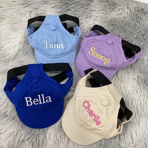 Op de afbeelding: Vier hondenbaseballpetten in blauw, paars, marineblauw en crème. Elke pet heeft een zwarte band en is geborduurd met de naam van een huisdier: "Luna", "Snoop", "Bella" en "Charlie".