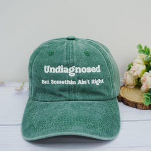 May include: A forest green baseball cap with a distressed, washed-out texture. The front features white embroidered text that reads "Undiagnosed" on the top line and "But Somethin Ain't Right" on the bottom line, centered above the curved brim.