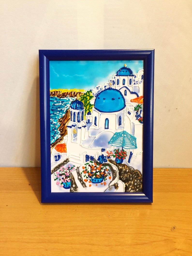 Puede incluir: Una pintura enmarcada que representa un edificio blanco con c&uacute;pulas azules, sobre un fondo de cielo azul y oc&eacute;ano. El edificio est&aacute; rodeado de flores y una sombrilla azul.