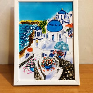 Puede incluir: Una obra de arte de vidrio emplomado enmarcada que representa un pueblo encalado con iglesias de c&uacute;pulas azules que dominan un mar turquesa. La escena est&aacute; llena de colores vibrantes y detalles intrincados, mostrando la belleza de una isla griega.