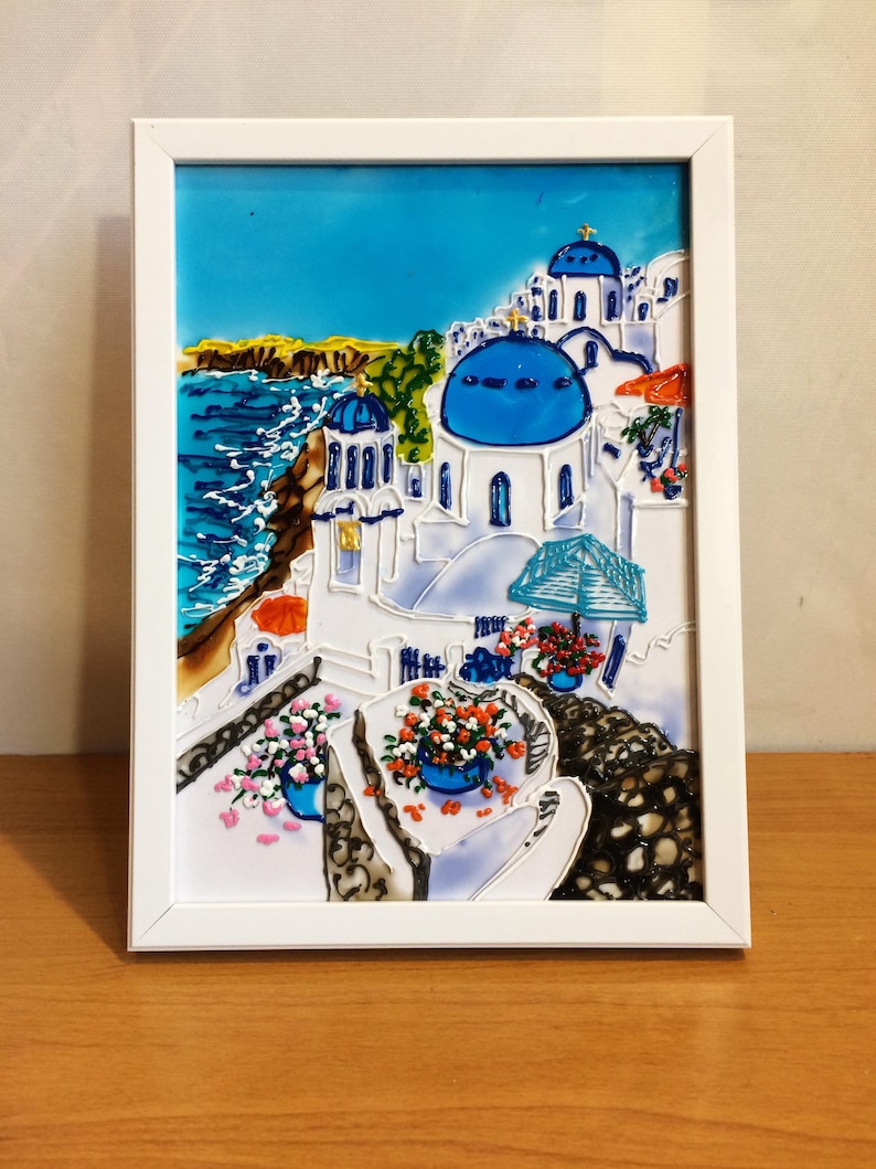 Puede incluir: Una obra de arte de vidrio emplomado enmarcada que representa un pueblo encalado con iglesias de c&uacute;pulas azules que dominan un mar turquesa. La escena est&aacute; llena de colores vibrantes y detalles intrincados, mostrando la belleza de una isla griega.