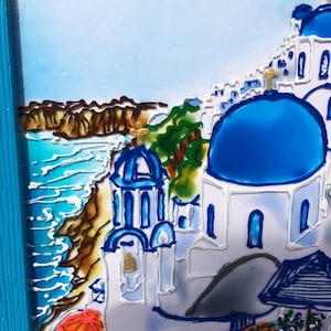 Puede incluir: Una pintura de un edificio blanco con un techo abovedado azul, sobre un fondo de mar azul y un acantilado rocoso. El edificio est&aacute; rodeado de otros edificios blancos y una colina verde. La pintura est&aacute; enmarcada en un marco azul.