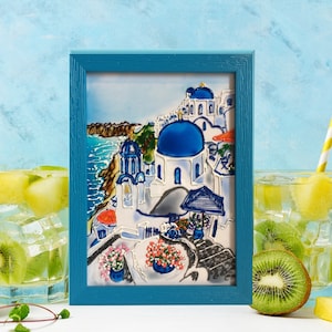 Puede incluir: Una pintura enmarcada de un pueblo griego blanco y azul con una iglesia de c&uacute;pula azul. La pintura est&aacute; sobre un fondo azul con un marco azul claro. Hay dos vasos de agua con fruta y pajitas delante de la pintura.