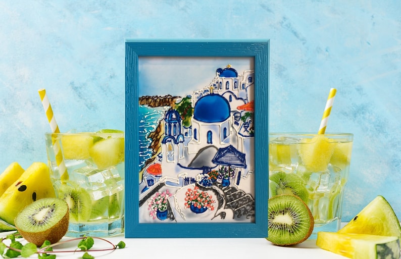 Puede incluir: Una pintura enmarcada de un pueblo griego blanco y azul con una iglesia de c&uacute;pula azul. La pintura est&aacute; sobre un fondo azul con un marco azul claro. Hay dos vasos de agua con fruta y pajitas delante de la pintura.