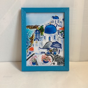 Puede incluir: Una pintura enmarcada de un pueblo griego blanco y azul con una iglesia de c&uacute;pula azul, edificios blancos y un mar azul en el fondo.