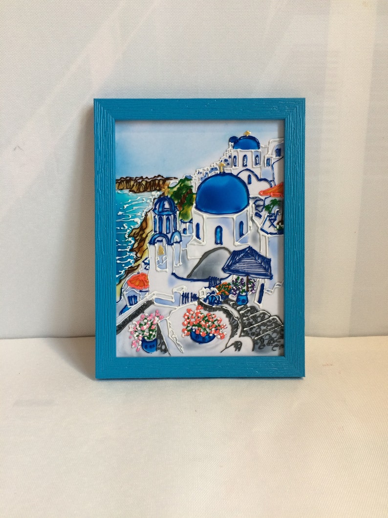 Puede incluir: Una pintura enmarcada de un pueblo griego blanco y azul con una iglesia de c&uacute;pula azul, edificios blancos y un mar azul en el fondo.