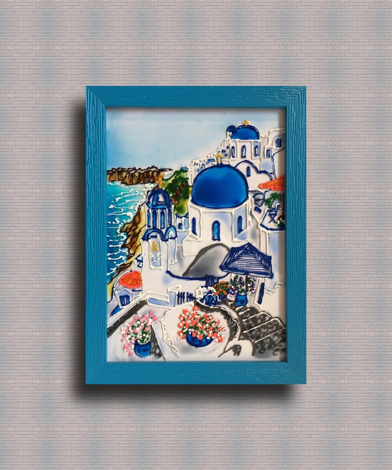 Puede incluir: Una pintura enmarcada de un pueblo griego blanco y azul con una iglesia de c&uacute;pula azul, con vistas a un mar azul. La pintura est&aacute; enmarcada en un marco azul turquesa.