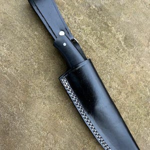 May include: A black leather knife sheath with a stitched edge. The sheath has a flap that secures with a snap.