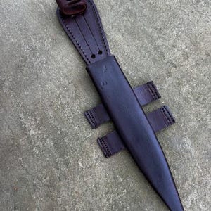 Puede incluir: Una funda de cuchillo de cuero marrón con cierre de presión metálico y trabilla para cinturón. La funda tiene una solapa que cubre la hoja del cuchillo.