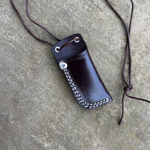 May include: A dark brown leather sheath with a decorative white stitched border and a silver-coloured rivet. The sheath is attached to a brown leather cord. The sheath is designed to hold a small object.