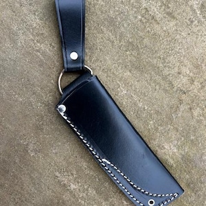 May include: A black leather knife sheath with white stitching and a belt loop. The sheath features a silver metal ring and rivets. The stitching follows the curved edges. The leather appears smooth and durable.