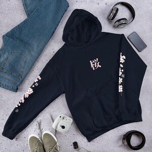 May include: A navy blue hoodie with a pink Japanese character on the front and a floral design on the sleeves. The floral design features pink blossoms and green leaves.
