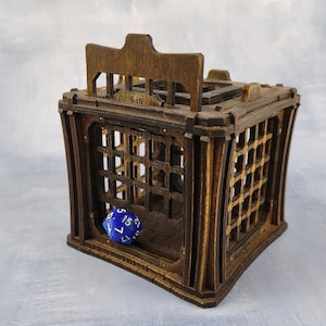 May include: A wooden dice jail with a cage design. The jail is made of dark brown wood and features a hinged top and barred sides. A blue, multi-sided die is inside. The word "fish" is etched on the top.