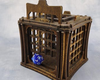 May include: A wooden dice jail with a cage design. The jail is made of dark brown wood and features a hinged top and barred sides. A blue, multi-sided die is inside. The word "fish" is etched on the top.