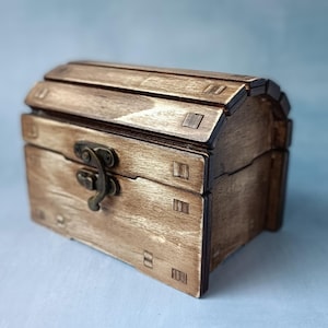 Op de afbeelding: Een kleine houten schatkist met een gebogen deksel. De kist is gemaakt van lichtbruin hout met donkerbruine accenten en een metalen sluiting. De kist is rechthoekig met decoratieve houten verbindingen.