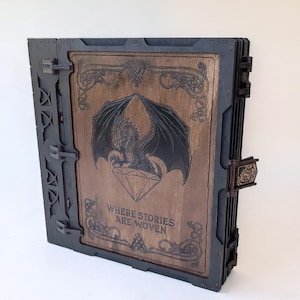 May include: A wooden book-shaped box with a dragon and diamond design on the front. The box is dark brown with black accents and the text "WHERE STORIES ARE WOVEN". The box has a clasp closure.