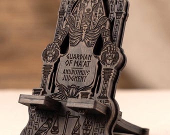 May include: A dark brown wooden phone stand featuring an intricate laser-engraved design of the Egyptian deity Anubis. The text reads GUARDIAN OF MA'AT ANUBISIMUIS' JUDGMENT, accompanied by hieroglyphic motifs, a scale, and a scarab beetle design.