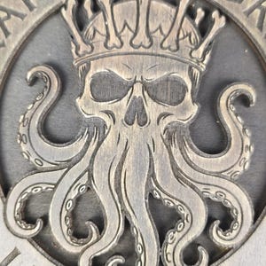 Puede incluir: Una placa circular de madera marrón oscuro con un diseño tallado de una calavera que lleva una corona hecha de huesos, con tentáculos de pulpo extendiéndose hacia abajo. El texto CAPTAIN'S LURK está tallado arriba y LIBERTALIA abajo.
