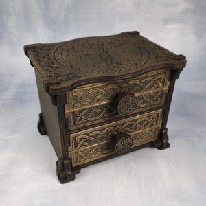 Op de afbeelding: Een donkerbruine houten kist met twee lades, voorzien van ingewikkelde Keltische knooppatronen op de ladefronten en een cirkelvormig boomontwerp op de bovenkant. Elke lade is uitgerust met een ronde knop met een triskelion-symbool.