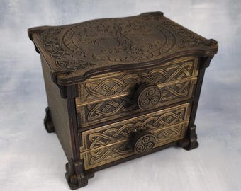 May include: A dark brown wooden chest with two drawers, featuring intricate Celtic knotwork patterns on the drawer fronts and a circular tree design on the top surface. Each drawer is equipped with a round knob displaying a triskelion symbol.