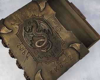 May include: A rectangular wooden box with a hinged lid featuring a raised dragon relief in the center. The surface has a weathered bronze finish with engraved runic symbols and four decorative corner accents. The interior is open and empty.