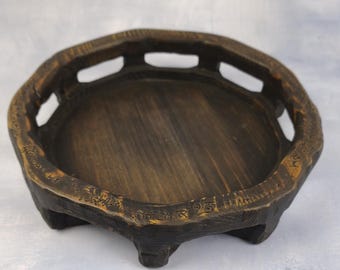 May include: A dark brown wooden display stand with a circular base and a raised, carved rim featuring cut-out sections. The wood has a visible grain texture with lighter brown highlights along the edges and is supported by four short, sturdy feet.