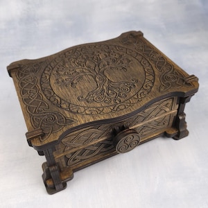 Può includere: Una scatola rettangolare in legno marrone scuro con un intricato disegno dell'albero della vita inciso al laser sul coperchio, circondato da motivi a intreccio celtico. Il pannello frontale include una manopola circolare con un triskelion.