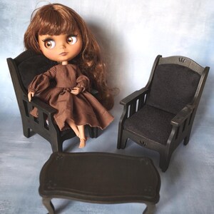 Op de afbeelding: Een pop met bruin haar en een bruine jurk zit in een miniatuur houten stoel. Een tweede stoel en een kleine tafel zijn ook zichtbaar. De meubels zijn donkerbruin met een matte afwerking en de pop heeft grote ogen.