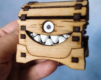 May include: A wooden box with a cartoonish monster face. The box is made of light brown wood with dark brown accents. It has a large, toothy grin and a single eye with a purple iris. The box is held in a hand.