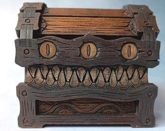 May include: A wooden treasure chest with a carved monster design. The chest features three eye-shaped cutouts and a row of teeth. The wood has a dark brown stain with lighter brown accents, and metal hinges and clasps.