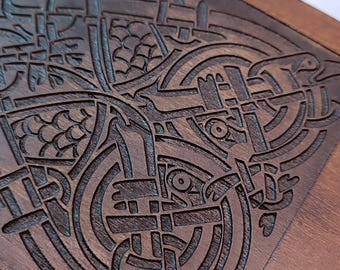 May include: Close-up of a wooden surface with a detailed, carved Celtic knot design. The dark brown wood features intricate patterns of interwoven lines, curves, and shapes, creating a textured, artistic appearance. The design is deeply etched into the wood.