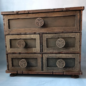 Può includere: Un cassettone in legno dall'aspetto rustico e artigianale. Il cassettone presenta quattro cassetti con pomelli circolari decorativi e un cassetto superiore. Il legno ha una finitura invecchiata, con venature visibili e colore marrone scuro.