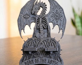 May include: A dark gray wooden phone stand featuring a skeletal dragon with outstretched wings. The wings are etched with eye symbols, runic patterns, and a compass design. The base displays the text BORN OF THE ABYSS in capital letters.