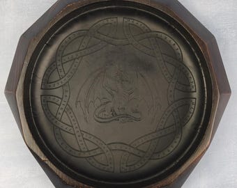 May include: A dark brown wooden tray with an octagonal outer shape and a circular interior. The center features an embossed dragon illustration surrounded by a circular band containing intricate, stylized runic-style lettering and symbols.