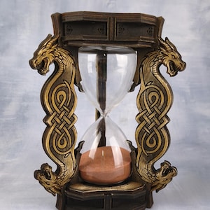 Könnte beinhalten: Sanduhr mit Holzrahmen, Drachen- und keltischen Knoten-Designs. Die klare Glaskugel enthält rotbraunen Sand. Der Rahmen ist dunkelbraun mit goldenen Akzenten.