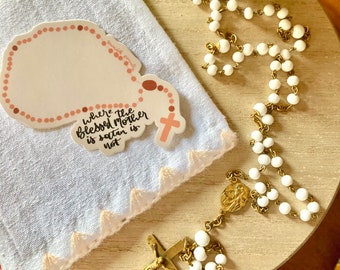 May include: A sticker with a rosary design and the text "Where the Blessed Mother is satan is not" is placed on a white cloth with a scalloped edge. A white beaded rosary with gold-colored metal accents is also present.