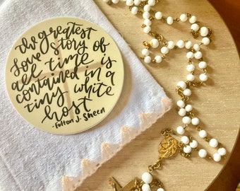 May include: A round, cream-colored sticker with the quote "the greatest story of all time is contained in a tiny white host" in black script. A white rosary with gold accents and a white cloth with a pink trim are also visible.