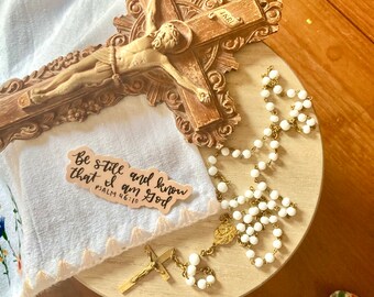 May include: A brown wooden crucifix with an INRI plaque, a white rosary with gold-toned metal links and a small crucifix, and a beige sticker featuring the black cursive text Be still and know that I am God PSALM 46:10 on a white cloth.