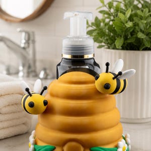 May include: A decorative soap dispenser featuring a yellow beehive design with two cartoon bees. The dispenser has a black pump with a gold accent and is decorated with green leaves and white flowers.
