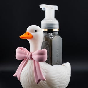 May include: A white duck-shaped soap dispenser holder with an orange beak and a pink bow. A clear soap dispenser with a white pump is placed in the holder. The background is black.