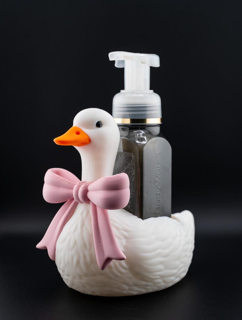 May include: A white duck-shaped soap dispenser holder with an orange beak and a pink bow. A clear soap dispenser with a white pump is placed in the holder. The background is black.